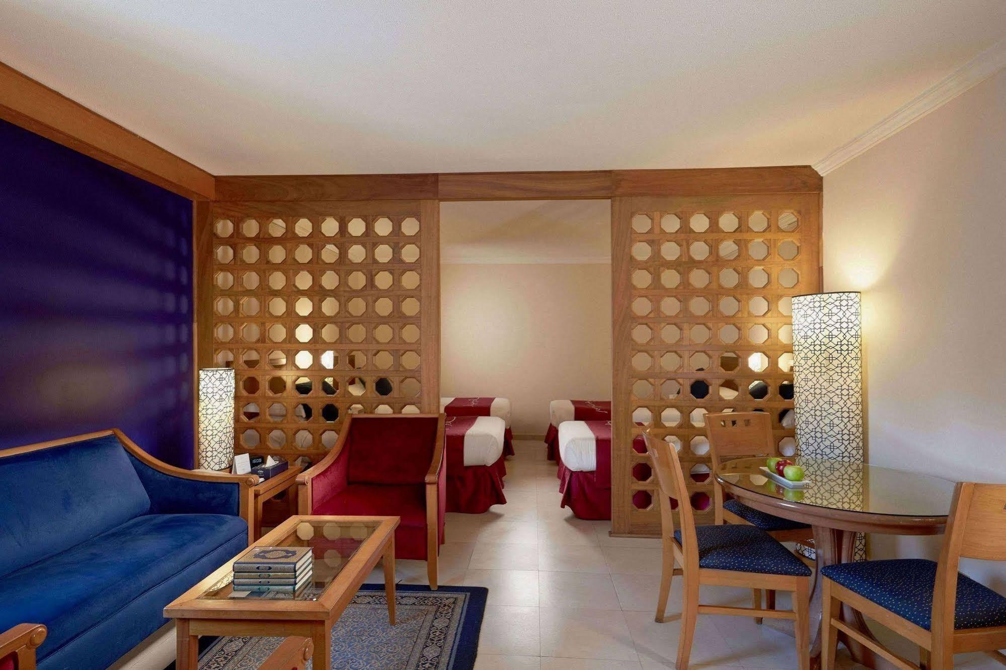 Makarem Ajyad Makkah Hotel Μέκκα Εξωτερικό φωτογραφία The photo shows a well-furnished hotel room or suite. On the left side, there is a seating area featuring a blue sofa and a small coffee table, as well as a couple of decorative items. The room is separated by a decorative wooden partition with circu