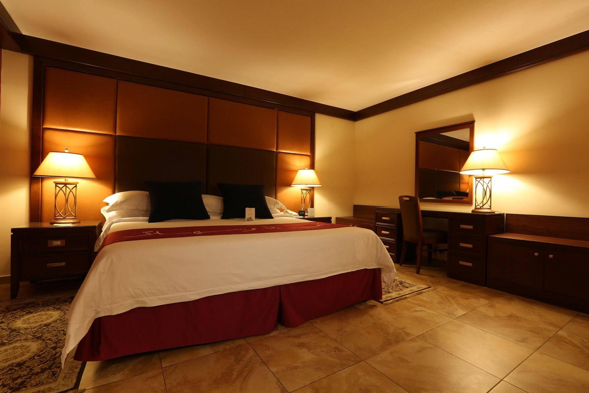 Makarem Ajyad Makkah Hotel Μέκκα Εξωτερικό φωτογραφία The photo shows a well-appointed hotel room with a cozy and inviting atmosphere. It features a large bed with a white comforter and dark pillows, positioned against a wooden headboard. There are two bedside lamps providing soft lighting on either sid