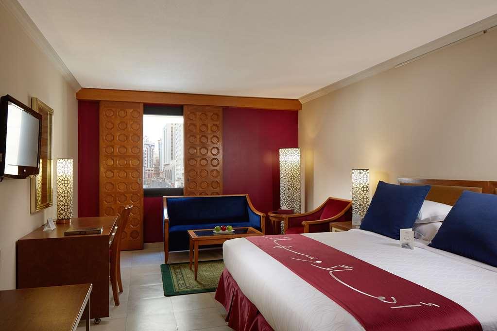 Makarem Ajyad Makkah Hotel Μέκκα Δωμάτιο φωτογραφία The photo showcases a hotel room featuring a modern design. In the center, there's a large bed with white linens and a decorative red runner. On either side of the bed, there are wooden nightstands with lamps. A blue sitting area is visible, composed