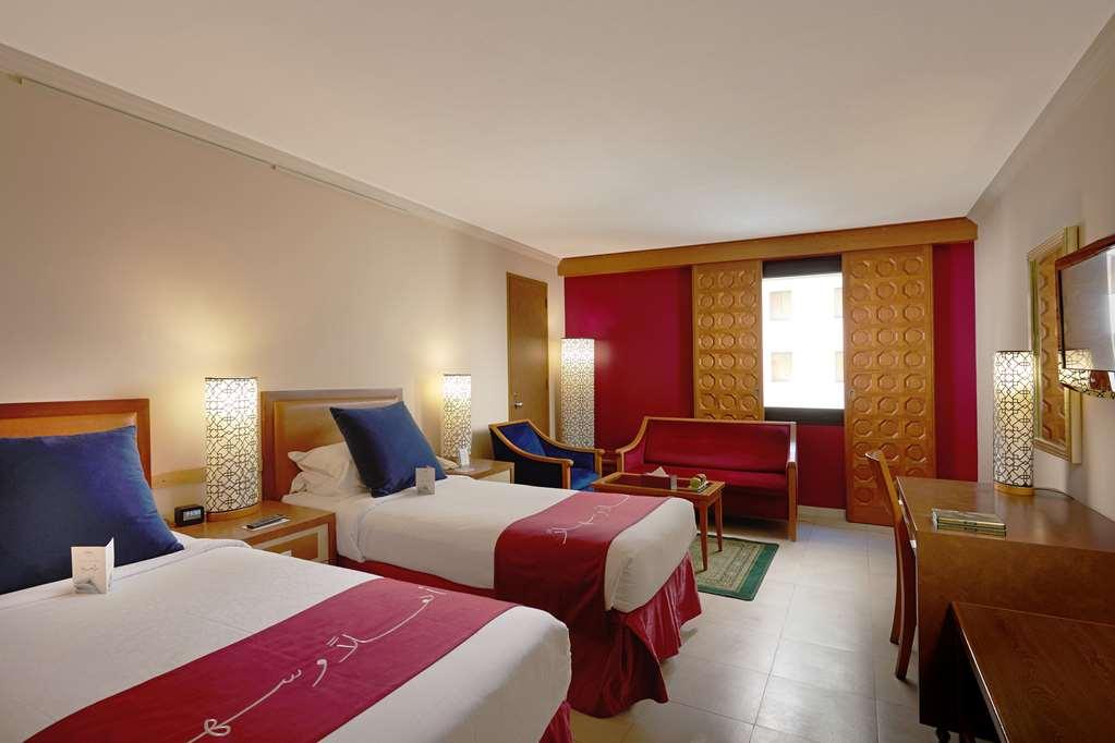 Makarem Ajyad Makkah Hotel Μέκκα Δωμάτιο φωτογραφία The photo shows a hotel room featuring two twin beds with red and white bedding. There are decorative lamps on either side of the beds. A small seating area is visible, including a blue chair and a red sofa. There is a wooden desk against one wall, a