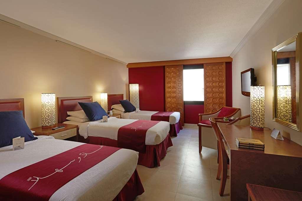 Makarem Ajyad Makkah Hotel Μέκκα Δωμάτιο φωτογραφία The photo shows a hotel room featuring three beds arranged with red and white bedding. The walls are painted in a neutral tone, and there are decorative lamps on nightstands beside each bed, providing ambient lighting. One of the walls has a window c