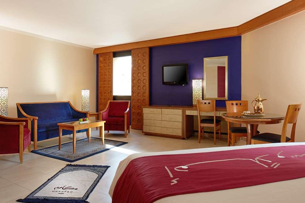 Makarem Ajyad Makkah Hotel Μέκκα Δωμάτιο φωτογραφία The image shows a hotel room with a modern, comfortable design. The room features a large bed with a red and white bedspread, a seating area that includes a blue sofa, two red chairs, and a wooden coffee table. There's a flat-screen TV mounted on the