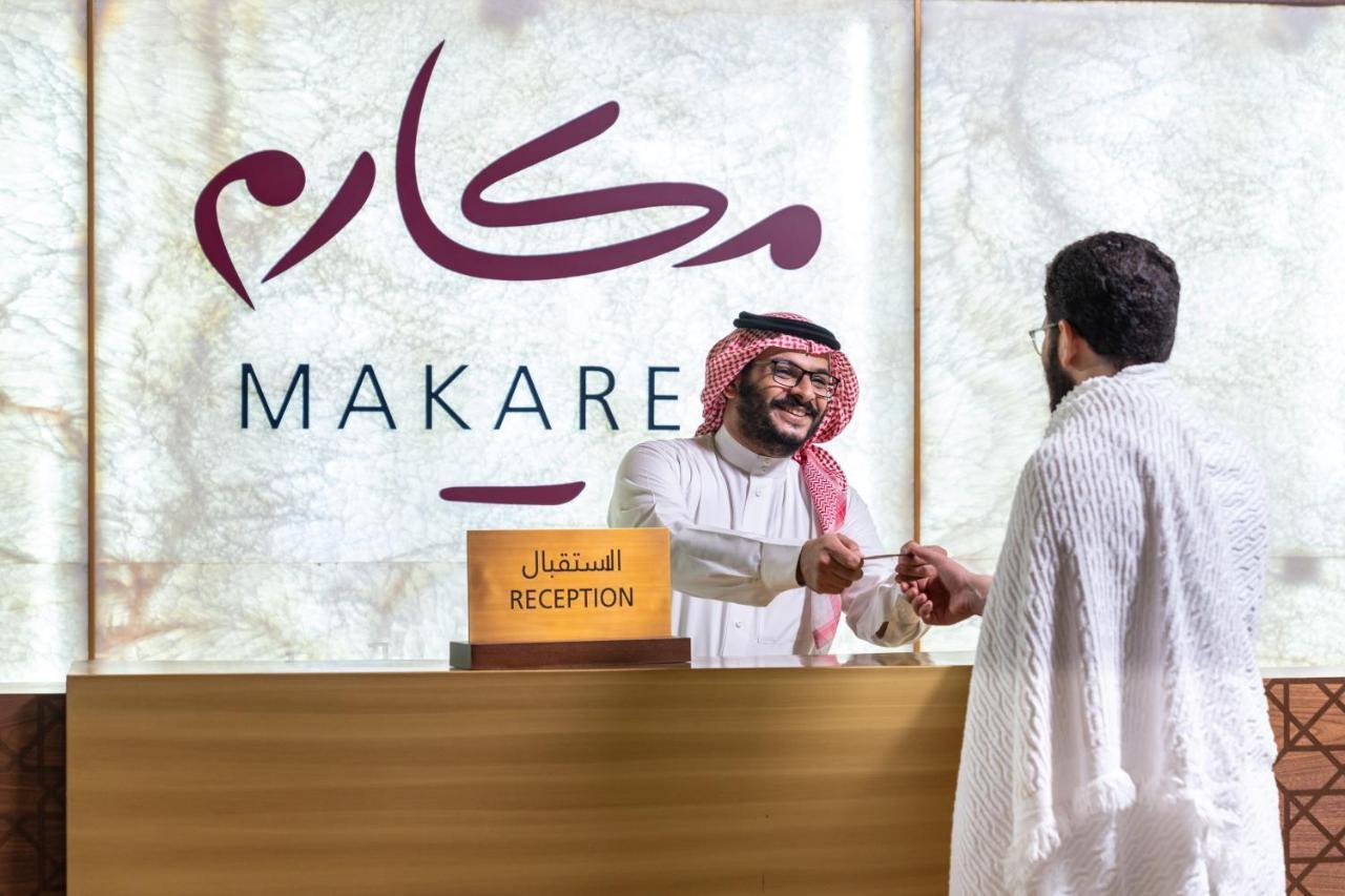 Makarem Ajyad Makkah Hotel Μέκκα Εξωτερικό φωτογραφία The photo shows a reception area with a sign that reads "MAKARE" in both Arabic and English. In the foreground, a staff member wearing traditional attire is smiling and shaking hands with a visitor, who is clad in a white garment, typical for formal 