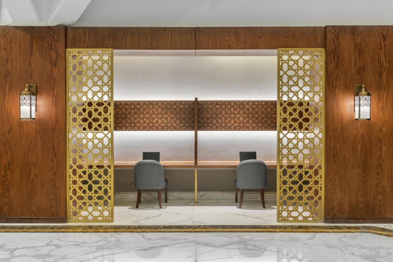Makarem Ajyad Makkah Hotel Μέκκα Εξωτερικό φωτογραφία The photo shows an elegantly designed space that features a reception or working area. There are two workstations with chairs positioned in front of them. The backdrop has a decorative panel with a geometric pattern. The area is framed by ornate gold