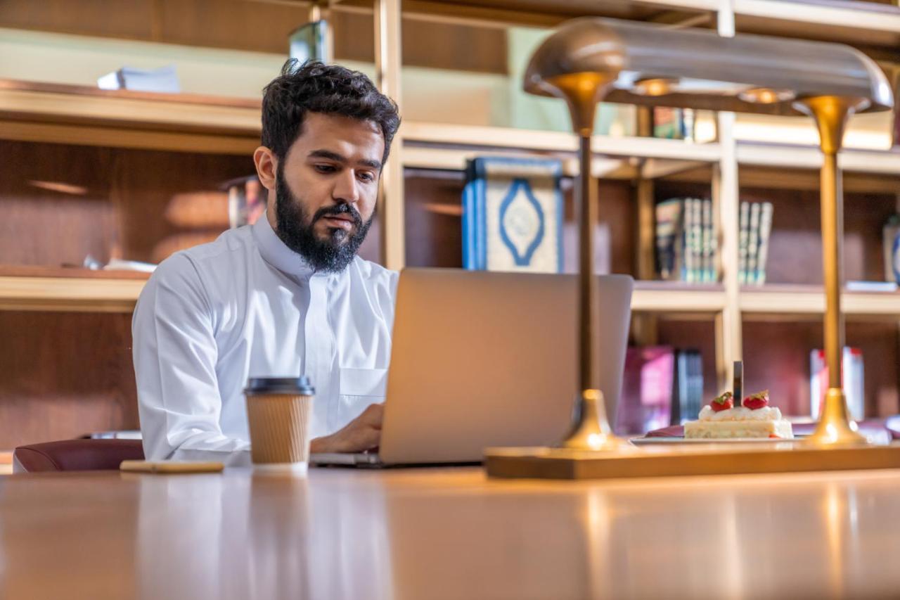 Makarem Ajyad Makkah Hotel Μέκκα Εξωτερικό φωτογραφία The photo shows a man sitting at a desk in a library or a study area. He is wearing a white traditional garment and is focused on working on a laptop. In front of him, there is a cup of coffee and a small cake decorated with strawberries. The backgro