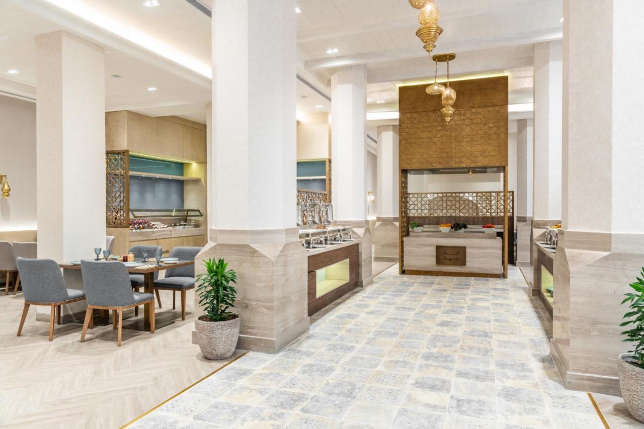 Makarem Ajyad Makkah Hotel Μέκκα Εξωτερικό φωτογραφία The photo depicts a modern, elegant dining area, likely inside a restaurant or hotel. It features a spacious layout with tall ceilings and large columns. The flooring is a light-colored tiled design. 

To one side, there are several dining tables and