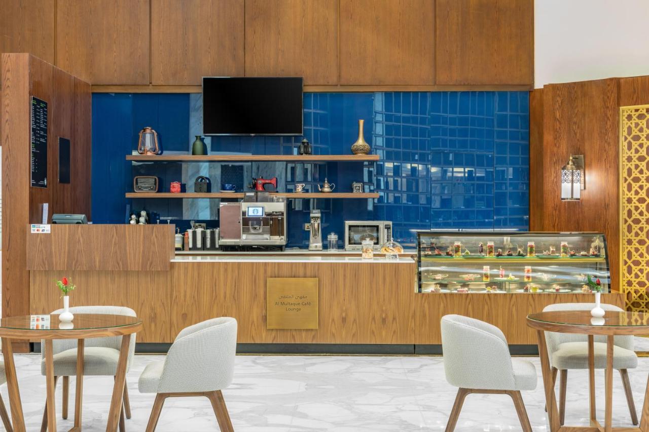 Makarem Ajyad Makkah Hotel Μέκκα Εξωτερικό φωτογραφία The photo shows the interior of a café. The design features warm wood paneling and a striking blue tiled wall. In the foreground, there are several light-colored chairs and small tables arranged neatly. Behind the counter, there's a variety of coffee