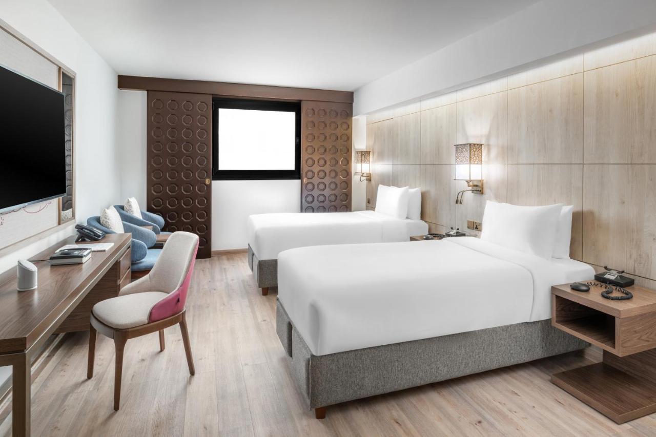 Makarem Ajyad Makkah Hotel Μέκκα Εξωτερικό φωτογραφία The photo shows a modern hotel room featuring two beds with white linens. There is a wooden accent wall with a window above it. Opposite the beds, a wall-mounted television is visible. A stylish wooden desk with a chair is situated on one side of the