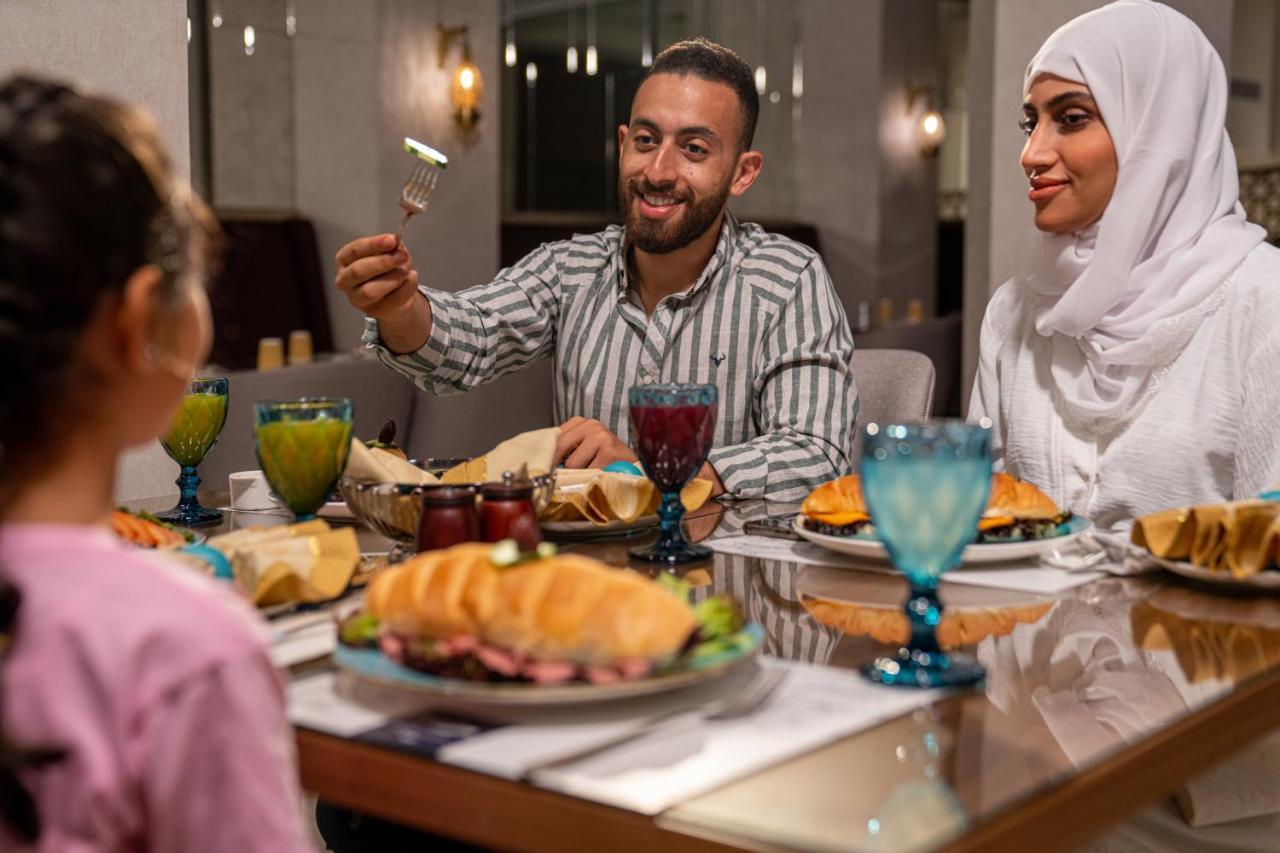 Makarem Ajyad Makkah Hotel Μέκκα Εξωτερικό φωτογραφία The photo shows a dining scene with three people enjoying a meal together. A man is seated at the table, smiling and holding a utensil, possibly ready to serve or share food. He is dressed in a striped shirt. Across from him is a woman wearing a whit