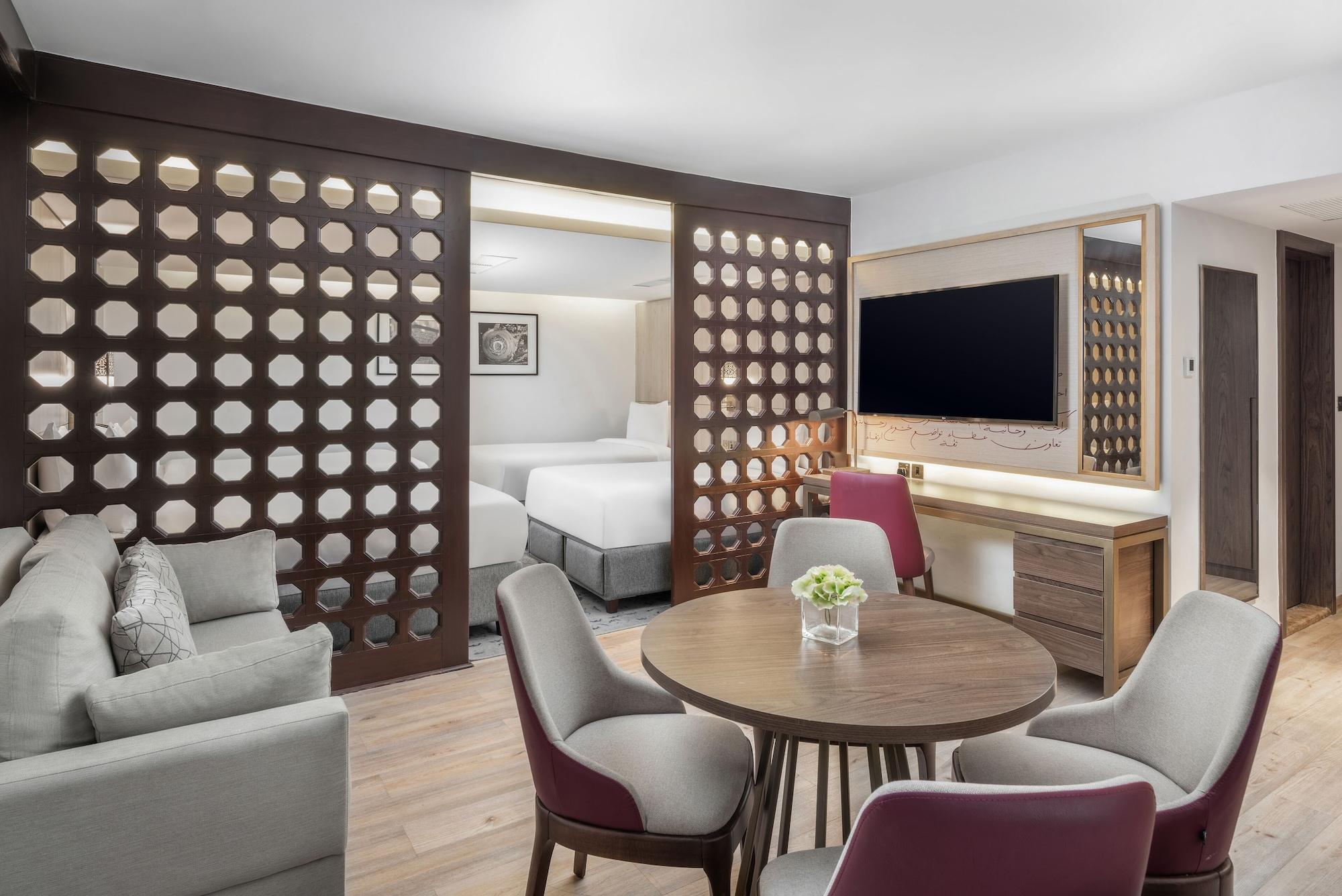 Makarem Ajyad Makkah Hotel Μέκκα Εξωτερικό φωτογραφία The photo shows a modern hotel room or suite. It features a living area with a comfortable gray sofa and a round wooden dining table surrounded by upholstered chairs. A large flat-screen TV is mounted on the wall opposite the seating area. There's a 