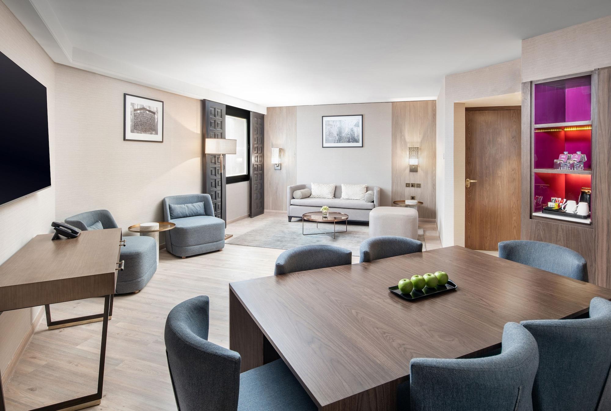 Makarem Ajyad Makkah Hotel Μέκκα Εξωτερικό φωτογραφία The photo shows a modern and elegantly furnished living area. It features a spacious layout with a comfortable seating arrangement that includes a sofa and armchairs. There is a large wooden dining table with several chairs around it. The walls are a