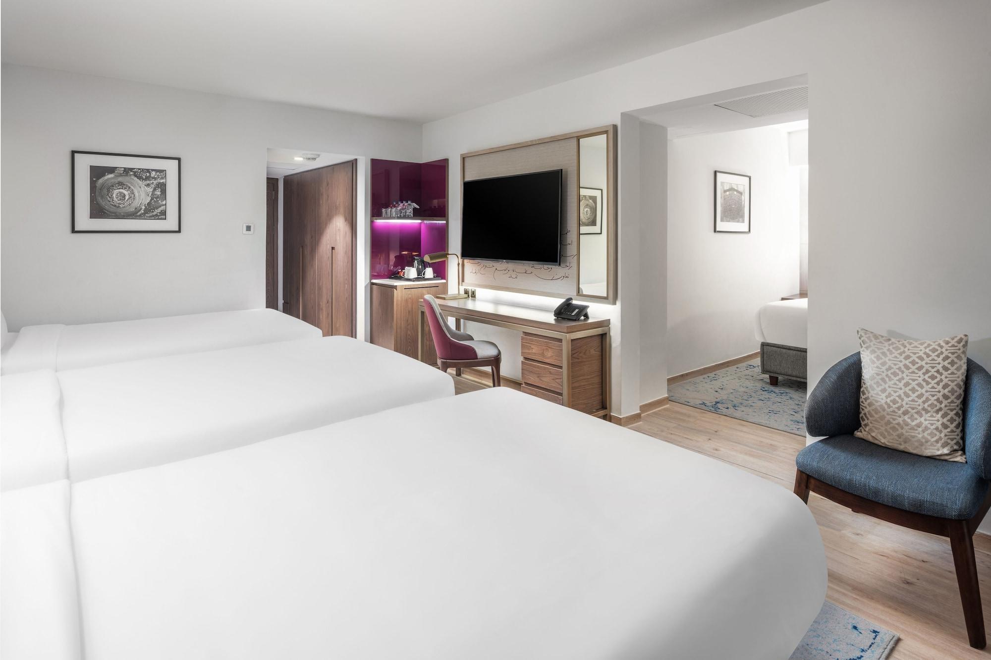 Makarem Ajyad Makkah Hotel Μέκκα Εξωτερικό φωτογραφία The photo shows a modern hotel room featuring two white beds with smooth linens. The room has a minimalistic design with light-colored walls and wooden flooring. On one side, there is a desk and chair next to a television mounted on the wall, along w
