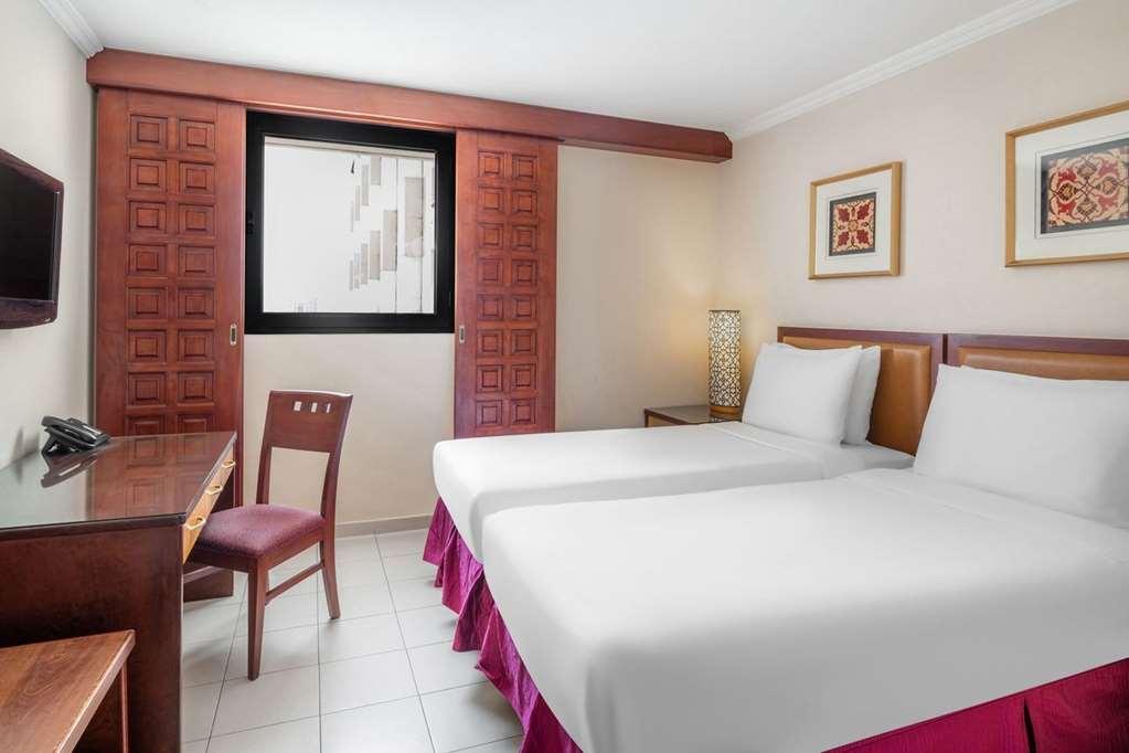 Makarem Ajyad Makkah Hotel Μέκκα Δωμάτιο φωτογραφία The photo shows a hotel room with two beds, each covered with white bedding. There is a wooden desk and a chair near the beds. The room features a window with a wooden frame and a decorative lamp beside the beds. On the wall, there are two framed art
