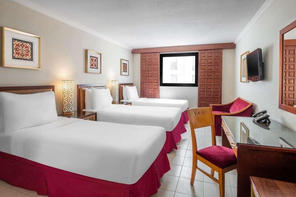 Makarem Ajyad Makkah Hotel Μέκκα Δωμάτιο φωτογραφία The photo shows a hotel room featuring two twin beds with white linens and red bed skirts. There are decorative elements on the walls, including framed artwork. A large window provides natural light, while a desk and a chair are positioned next to it