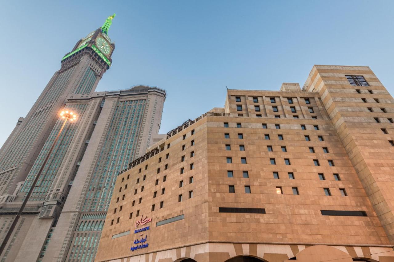 Makarem Ajyad Makkah Hotel Μέκκα Εξωτερικό φωτογραφία The photo shows a prominent building with a modern architectural design, likely a hotel or commercial establishment, at the forefront. It features a light-colored stone facade with large windows. Behind it, there is a tall skyscraper, which has a dis