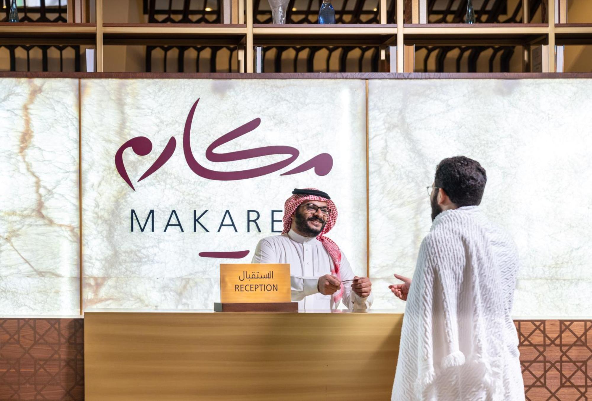 Makarem Ajyad Makkah Hotel Μέκκα Εξωτερικό φωτογραφία The photo depicts a reception area in a hotel or establishment named "Makarem." In the image, a receptionist, dressed in traditional attire, is smiling and interacting with a guest who is wearing a white robe, commonly known as a thobe or ihram. The 