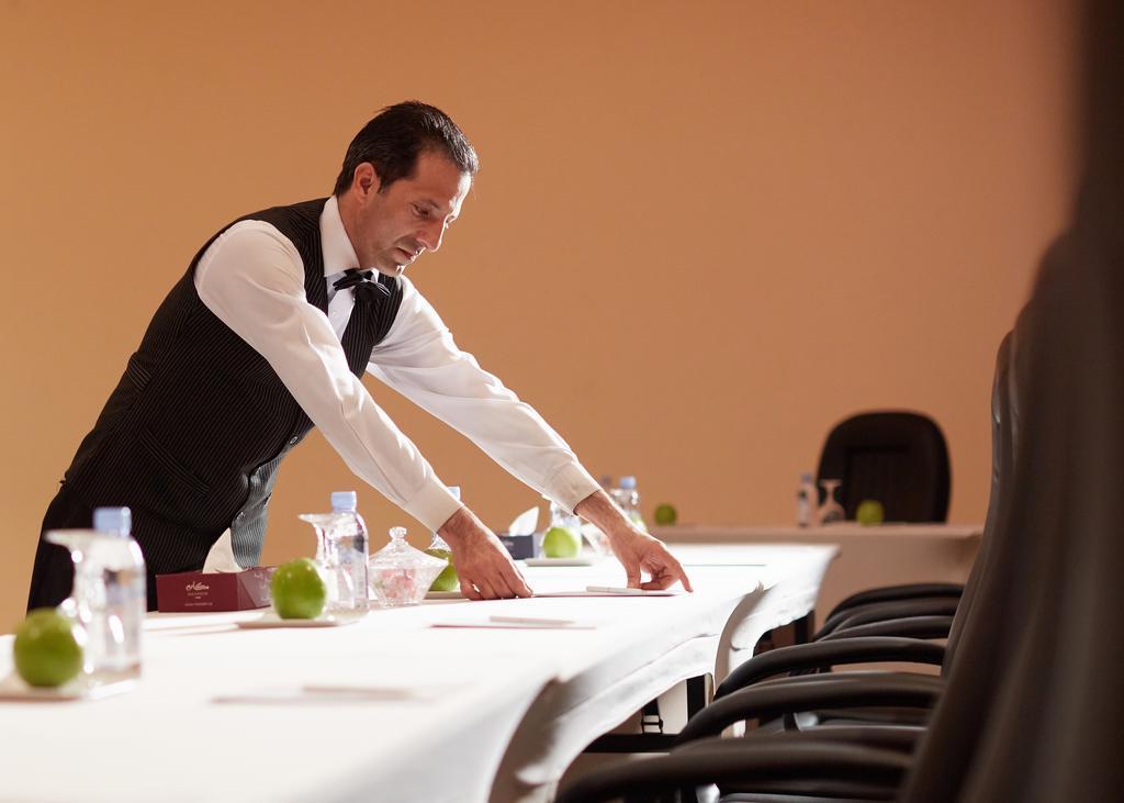 Makarem Ajyad Makkah Hotel Μέκκα Εξωτερικό φωτογραφία The image shows a waiter or service staff member in a formal attire, which includes a vest and bow tie. He is preparing a long table, likely for an event or meeting. The table is set with items such as water bottles and green apples, and there are se