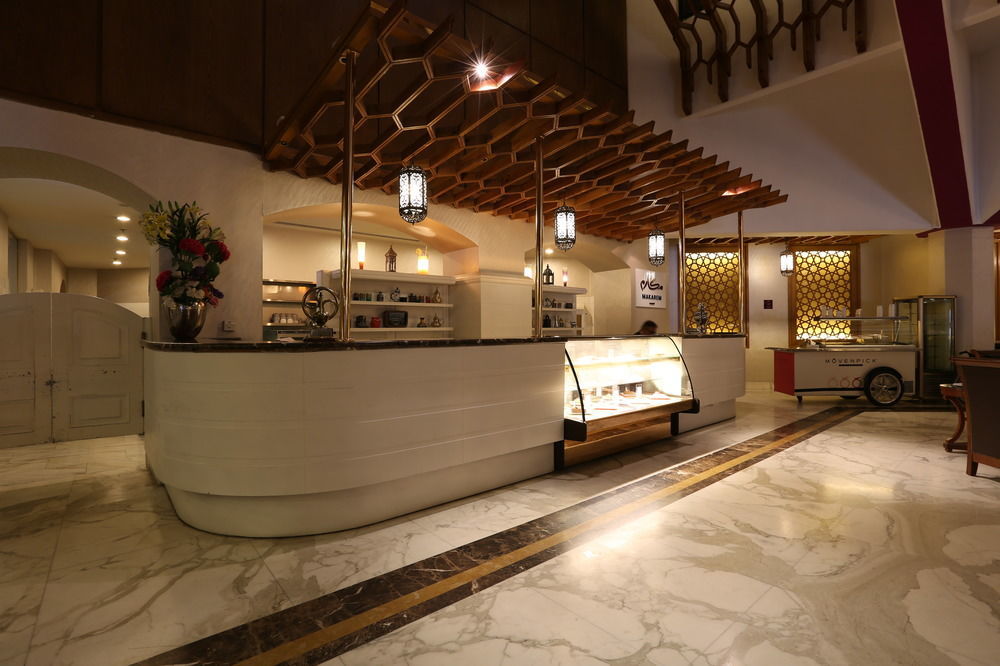Makarem Ajyad Makkah Hotel Μέκκα Εξωτερικό φωτογραφία The photo shows a modern reception area or lobby of a hotel or restaurant. The focal point is a curved front desk made of white material, featuring a glass display case that showcases various items. Above the desk, there is a decorative wooden struct